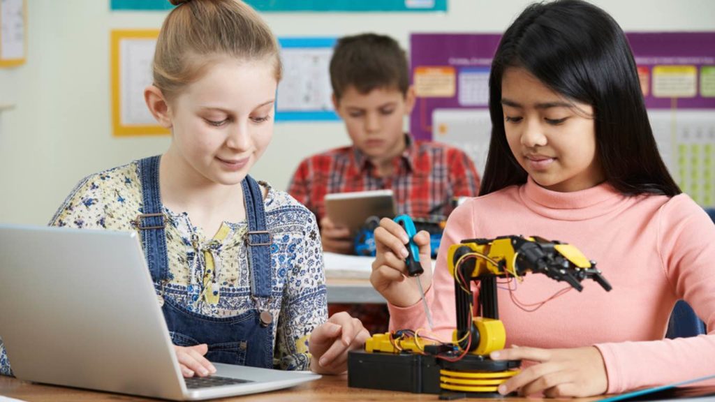 2 students fixing a robotic arm, while a third student is working on his iPad in the background